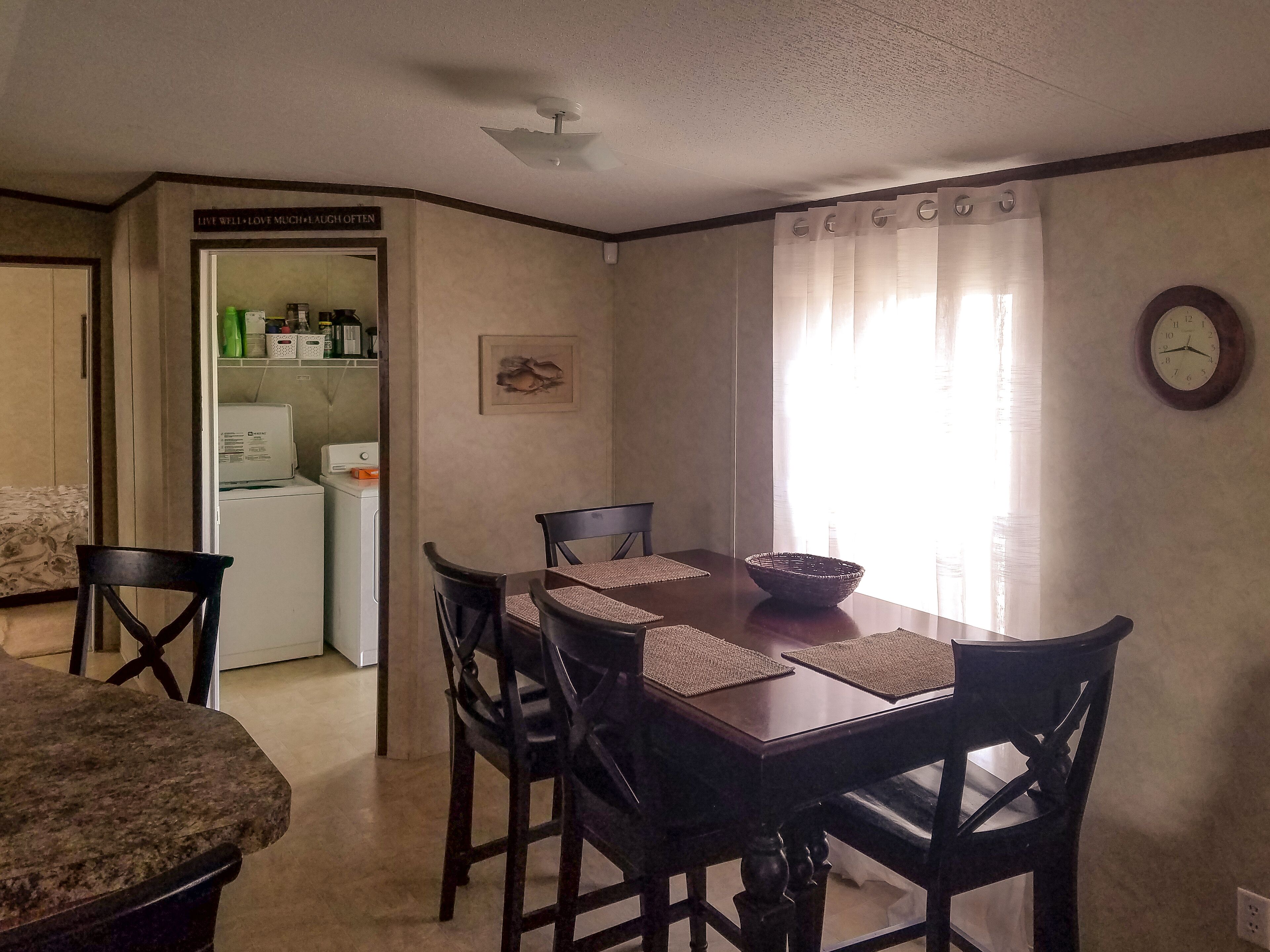 A dining area with a wooden table, four chairs, and a woven bowl. A clock and a framed picture adorn the walls.