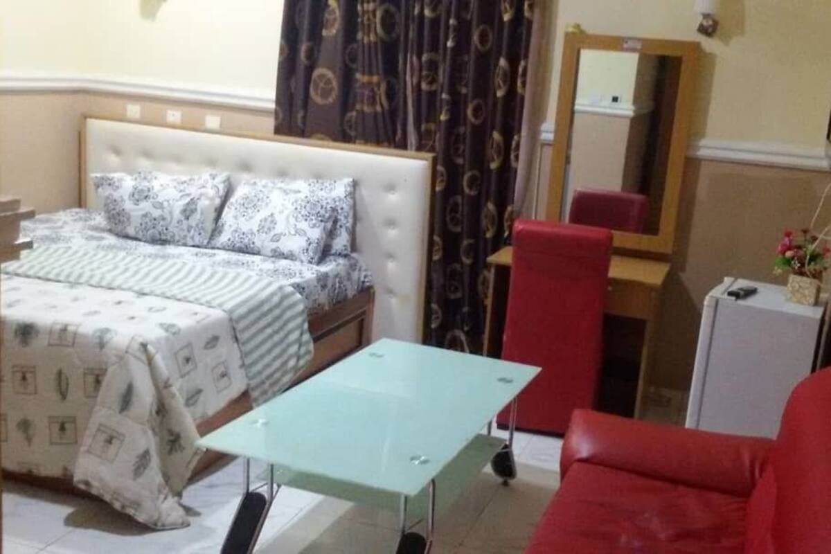 Quarto de hotel com cama, mesa pequena, cadeira vermelha, escrivaninha e espelho.
