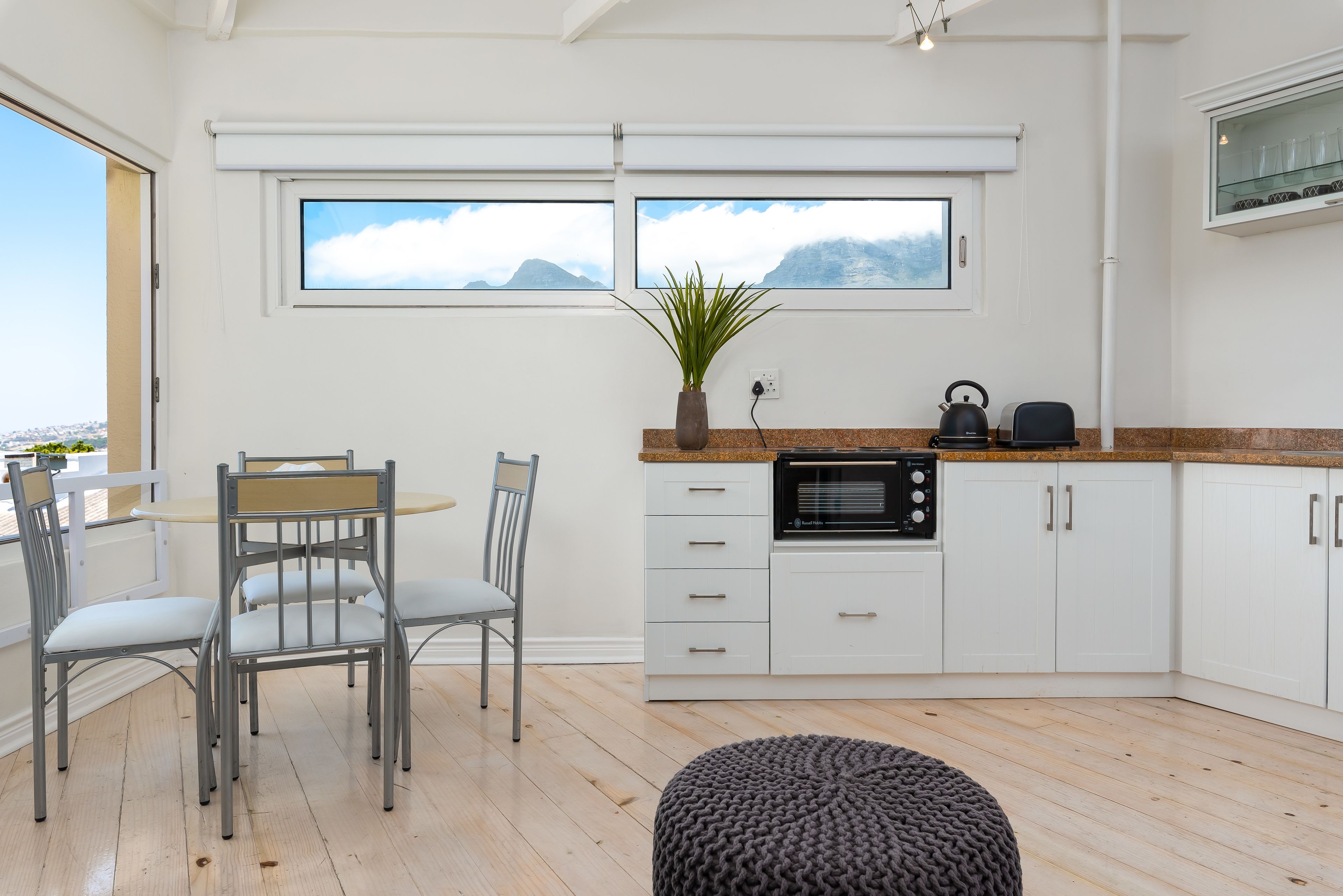 A modern kitchen with white cabinets, a dining table with chairs, a large window with a view of mountains, and a small plant on the counter.