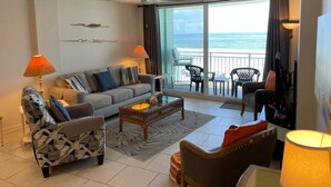 Una sala de estar con sofá, sillón y mesa de centro, un balcón con sillas y vista al océano.