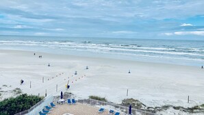Una playa con vista despejada al océano, un camino de arena que lleva al agua y una sección de la playa con sillas de descanso azules y sombrillas.