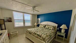 Un dormitorio con cama, ventilador de techo, ventana con persianas, mesita de noche con lámpara y cómoda.