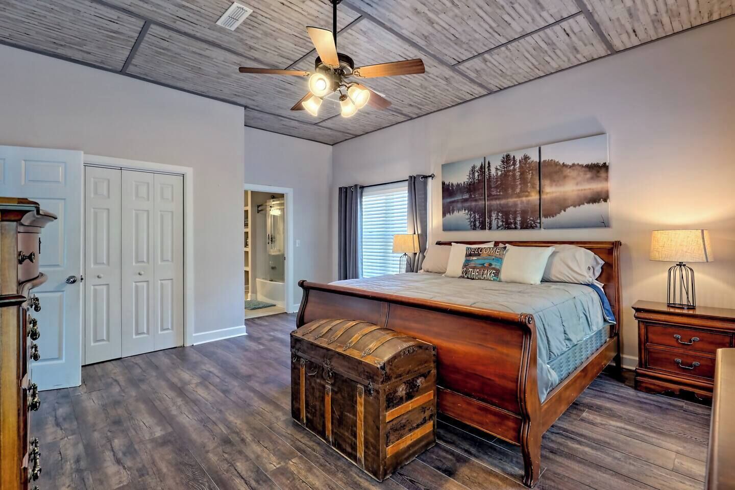 Une chambre avec un lit en bois, un ventilateur de plafond, une grande fenêtre avec des stores, un tableau représentant une forêt et une commode en bois.