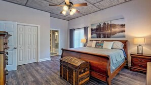 A bedroom with a wooden bed, a ceiling fan, a large window with blinds, a painting of a forest, and a wooden chest. - Lake Chatuge Bungalow by Chatuge Home Concierge | Cozy Pet-Friendly Retreat · Lake & Mountain Views · Firepit (Hiawassee)