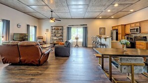 A spacious living room with a leather sofa, a coffee table, and a bookshelf. There is a dining area with a wooden table and chairs. The kitchen features modern appliances and wooden cabinets. - Lake Chatuge Bungalow by Chatuge Home Concierge | Cozy Pet-Friendly Retreat · Lake & Mountain Views · Firepit (Hiawassee)