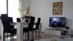 Un salon moderne avec une table basse en verre, des chaises noires, un meuble TV blanc et une plante en pot.