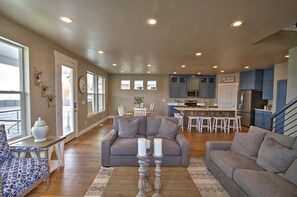 A spacious living room with a gray sofa, a patterned chair, and a coffee table with candles. A dining area with a white table and chairs is visible in the background. - Barefoot Bungalow #9 - Pool, Hot Tub, and Lake Access (Garden City)