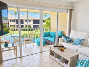 A modern living room with a glass door leading to a pool area, a white sofa with patterned cushions, and a small coffee table with books.