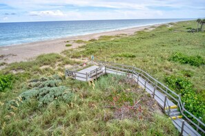 A beach with a wooden boardwalk leading to a small wooden structure, surrounded by grass and shrubs.