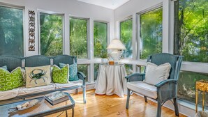 A spacious room with large windows, a sofa with patterned cushions, a table with a lamp, and a chair.
