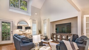 A spacious living room with a high ceiling, a large flat-screen TV, two dark sofas, a round coffee table, and a dining area with a white table and chairs.