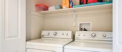 A laundry room with two white washers and dryers, a shelf with laundry supplies, and a door leading to another room.