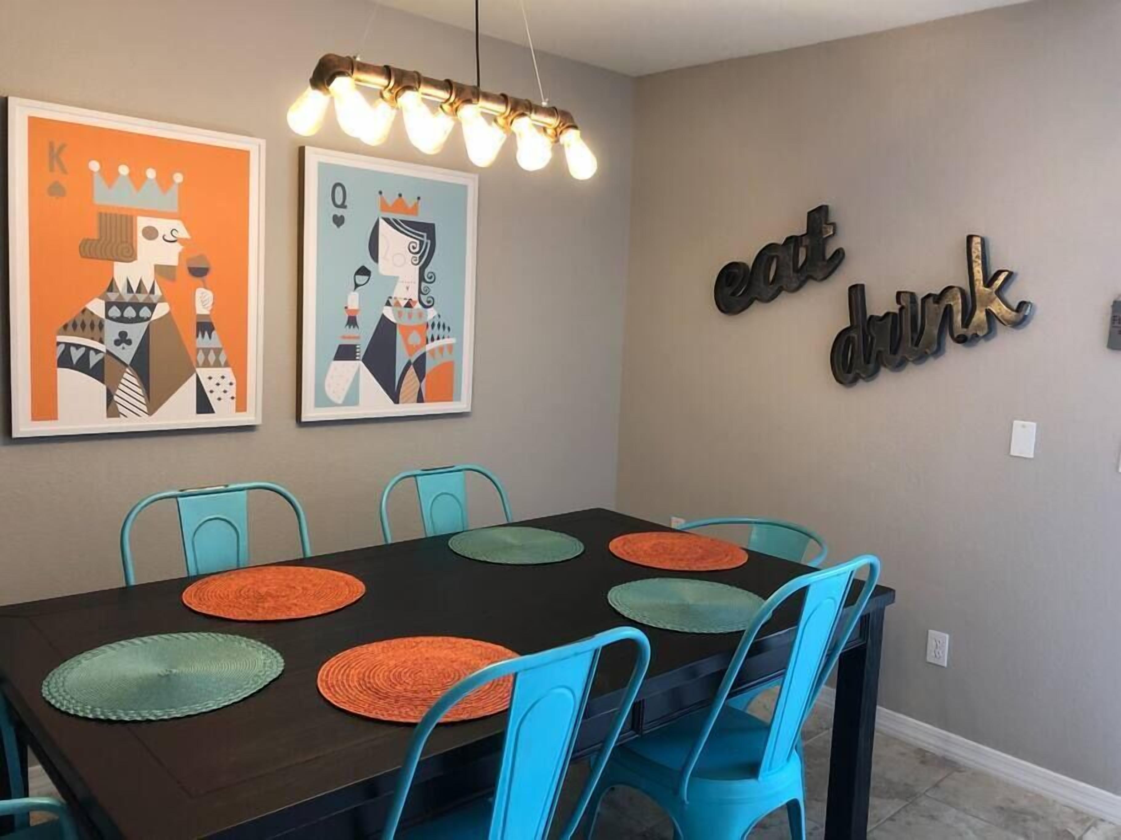 Ein Essbereich mit einem dunklen Holztisch, blauen Stühlen, orangefarbenen Unterlagen und zwei gerahmten Kunstwerken an der Wand.