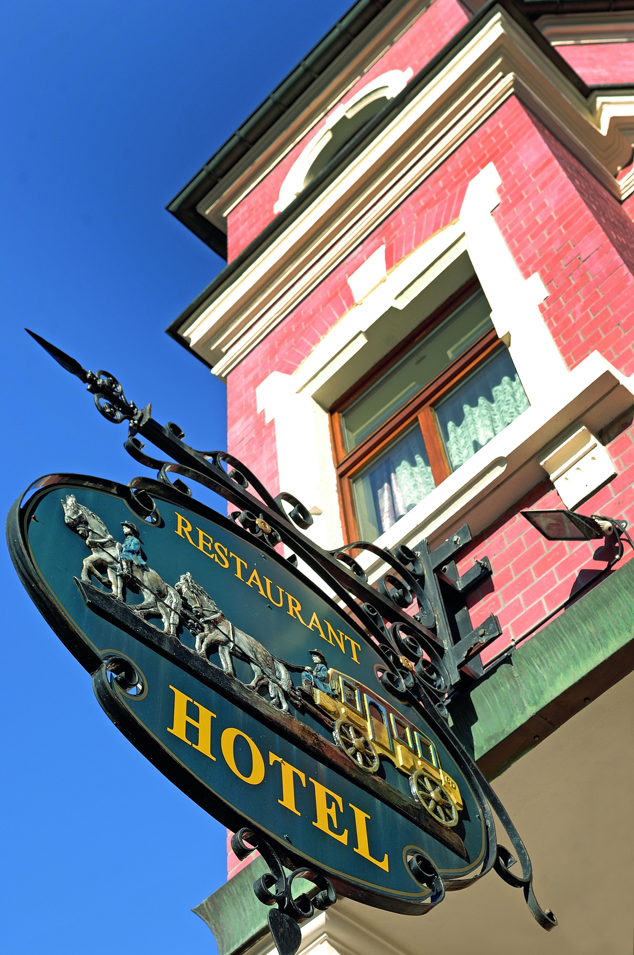 Ein Hotel Schild mit einer Illustration von Pferden und einer Kutsche, darüber ein Restaurantschild und im Hintergrund ein rot backsteins Gebäude.