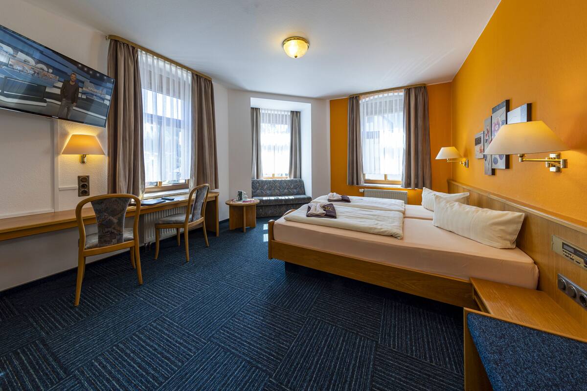 A hotel room with a large bed, a desk, a chair, a television, and two windows with curtains.