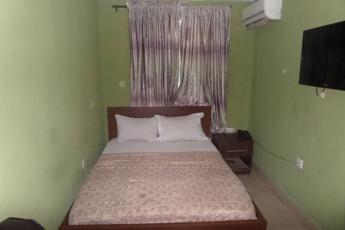 A small, single-bed room with a wooden headboard, a bedside table, a power outlet, and a wall-mounted air conditioner.