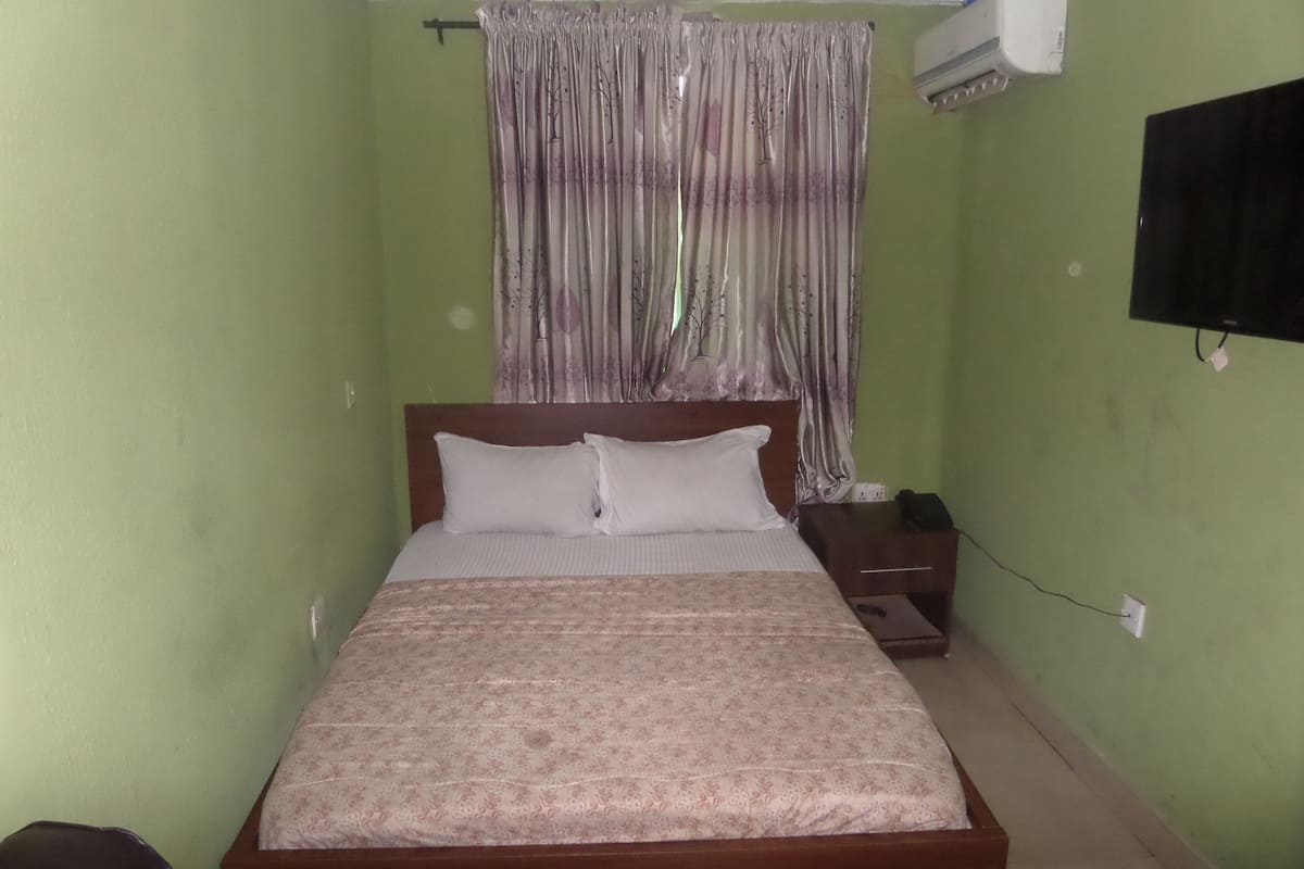 A bedroom with a bed, a nightstand, a wall-mounted TV, and a window with curtains.