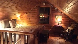 A cozy attic bedroom with a wooden ceiling, a bed with pillows, a rocking chair, and two bedside lamps.