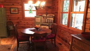 A wooden cabin interior with a round dining table, chairs, a stove, and a window with a view of trees.