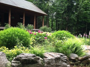 A rustic lodge with a covered porch, surrounded by lush greenery and flowering plants. - Perry Pond Paradise! (Narrowsburg)