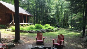 A cabin with a fire pit and two Adirondack chairs in a forest setting.