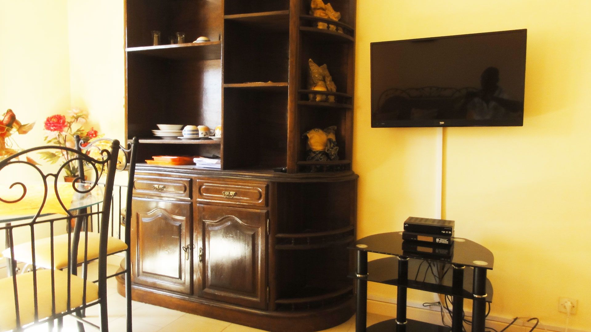 A corner room with a dark wooden cabinet, a small round table with a device, and a TV mounted on the wall.