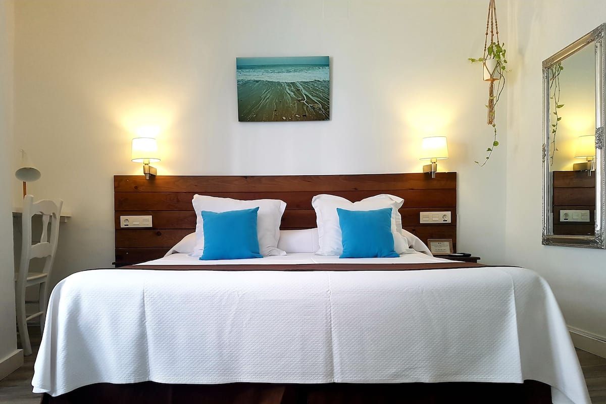 A double bed with white bedding and two blue pillows, a wooden headboard, a wall-mounted painting of a beach, and two wall-mounted lamps.