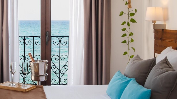 A hotel room with a bed, a bedside table with glasses and a bottle, a view of the sea through a balcony door, and a plant on the wall. - Hostal Casa del Mar (Altea)