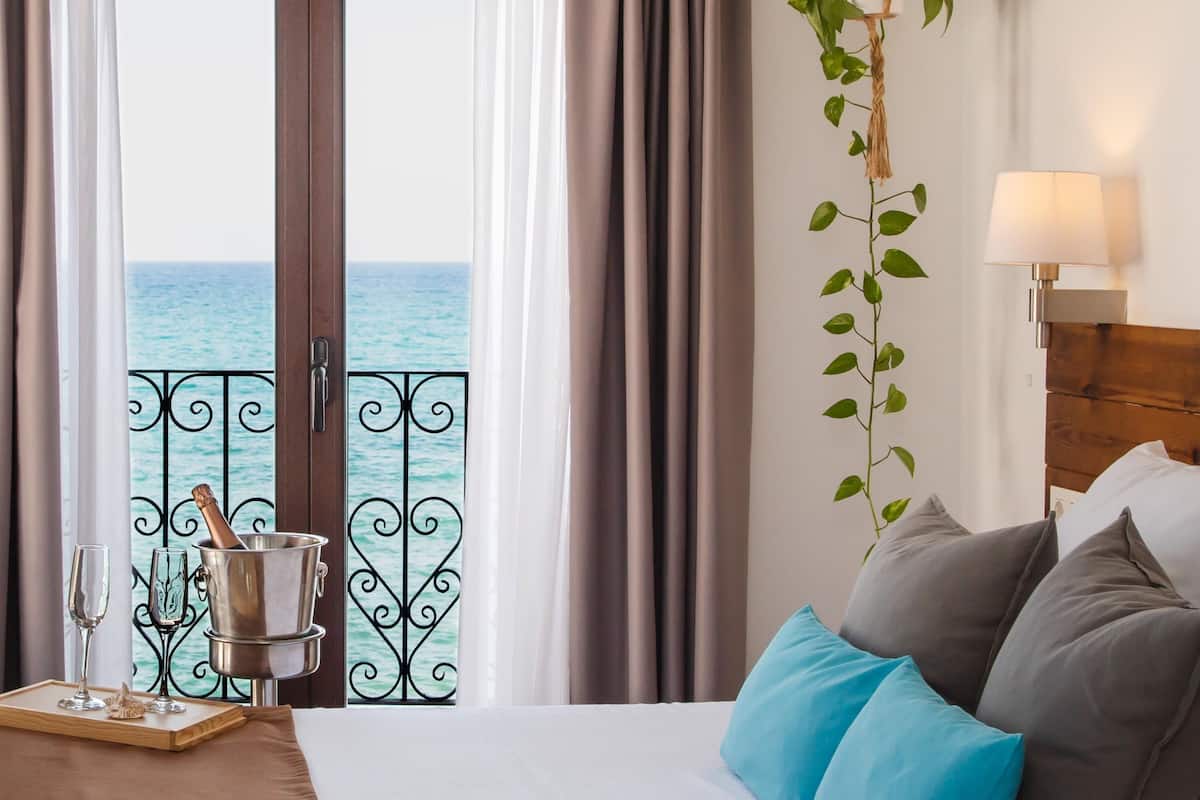 A hotel room with a bed, a bedside table with glasses and a bottle, a view of the sea through a balcony door, and a plant on the wall.