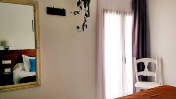 A room with a bed, a chair, a mirror, a window with curtains, and a wall-mounted plant.