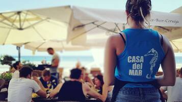 A person with a blue tank top and jeans at a seaside restaurant with outdoor seating and umbrellas.