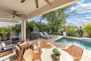 A covered patio with outdoor furniture, a pool, and lush greenery.