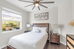 A bedroom with a bed, a ceiling fan, a nightstand, a lamp, a window with a view of foliage, and a painting on the wall.