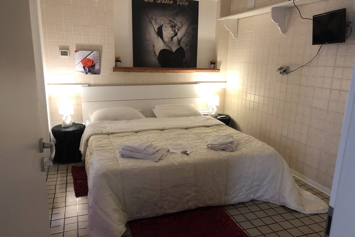 Habitació d'hotel amb un llit, dues taules de nit, un televisor penjat a la paret i un cartell.