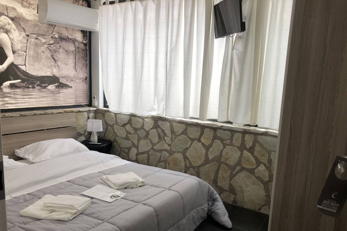 Un llit ben fet amb un edredó gris, una paret de pedra, una finestra amb cortines blanques i un televisor penjat.