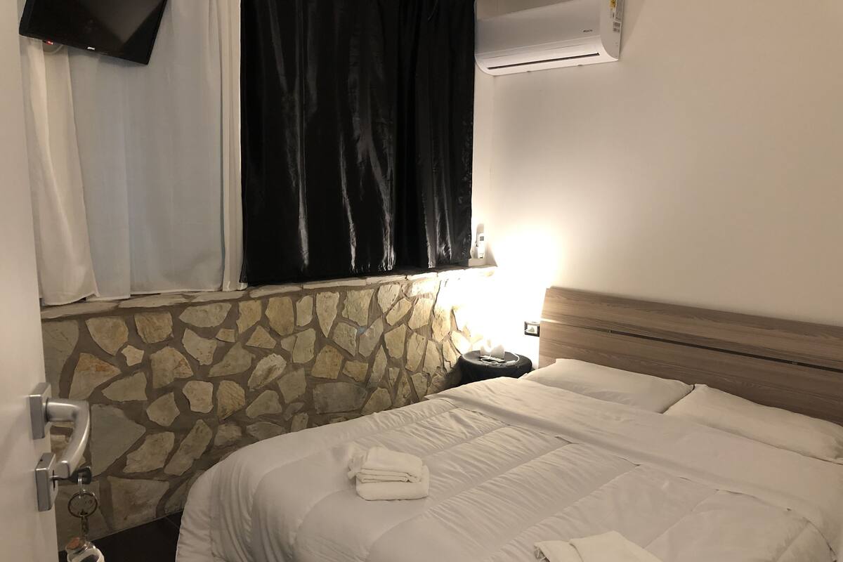 Habitació d'hotel amb paret de pedra, llit amb roba de llit blanca, tauleta de nit amb làmpada i televisió penjada a la paret.