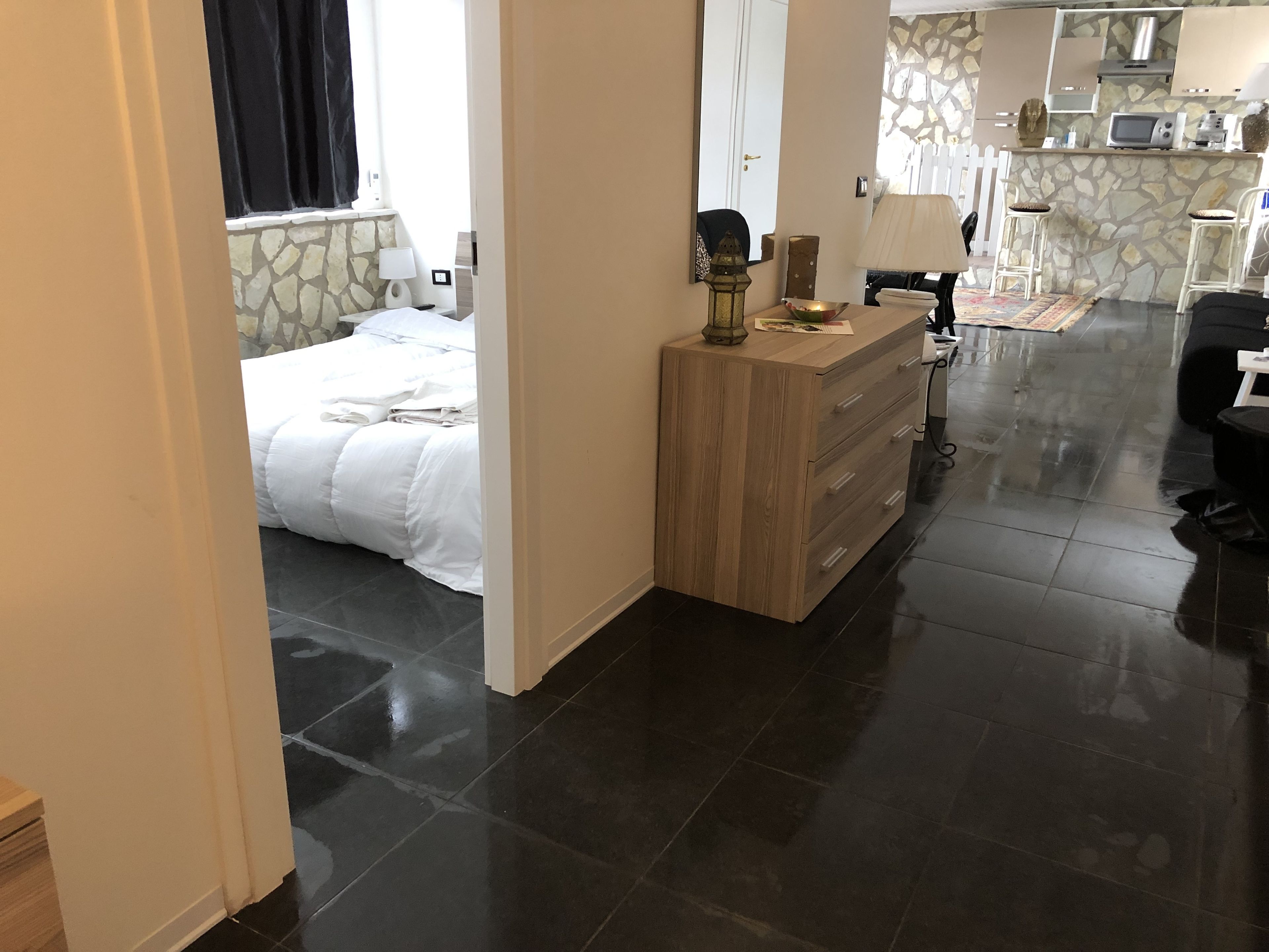 A hotel room with a bed, a wooden dresser, a small kitchen area with a microwave, and a stone wall.