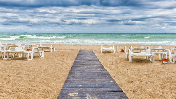 Una playa con un paseo de madera que lleva a sillas y mesas de plástico blanco, con vista al océano.