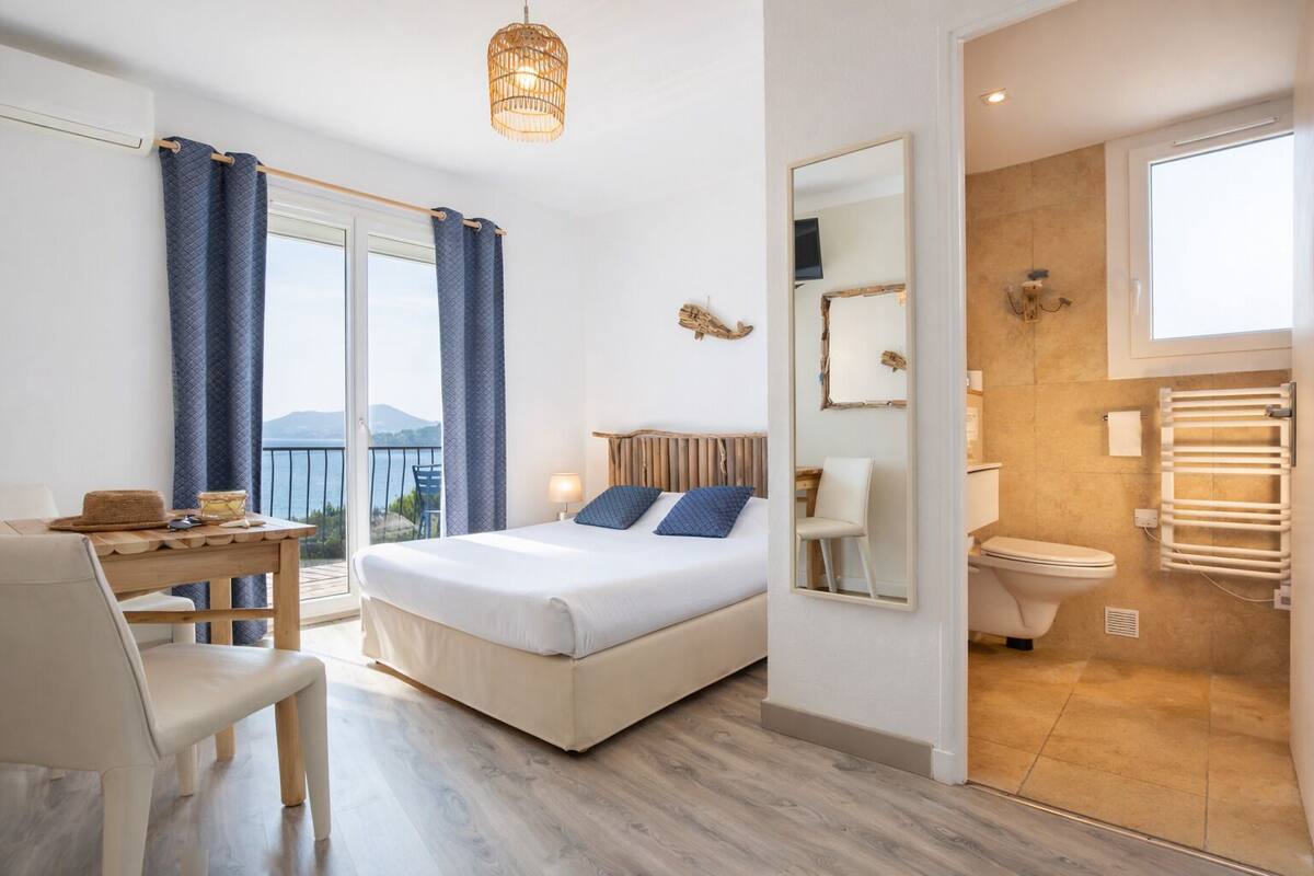 Quarto de hotel com cama, escrivaninha, cadeira, varanda com cortinas, banheiro com vaso sanitário e espelho.