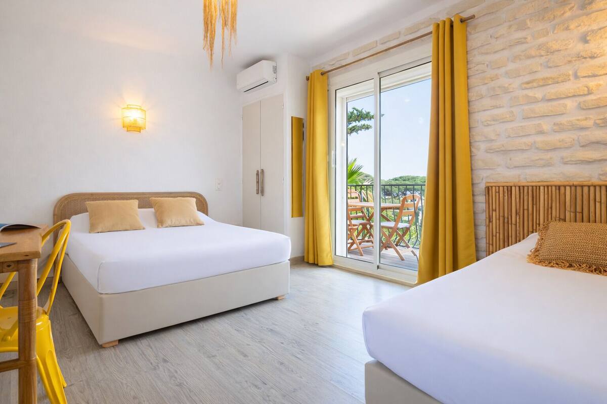 Quarto de hotel com cama, escrivaninha, cadeira, armário e varanda com cortinas amarelas.