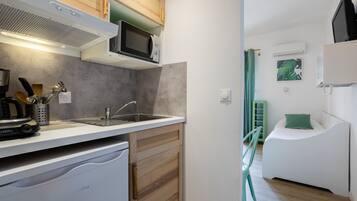 A compact kitchen with a microwave, sink, and coffee maker. There's a small refrigerator and wooden cabinets. A green chair is visible in the background.