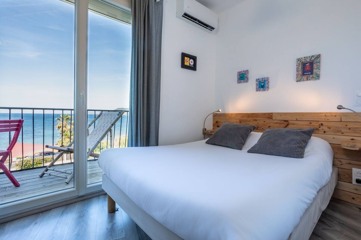 Quarto com uma cama grande, cabeceira de madeira e vista para o mar através de uma varanda.