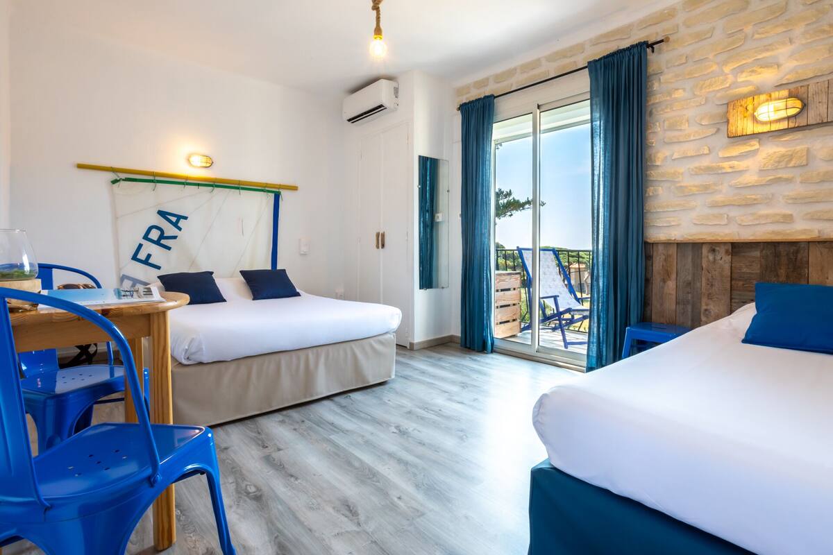 Quarto de hotel com duas camas, uma mesa com cadeira, uma janela grande com cortinas e uma parede de pedra.
