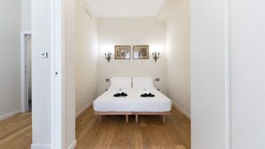 Une chambre avec un plancher en bois, un lit blanc, deux appliques murales et deux tableaux encadrés.