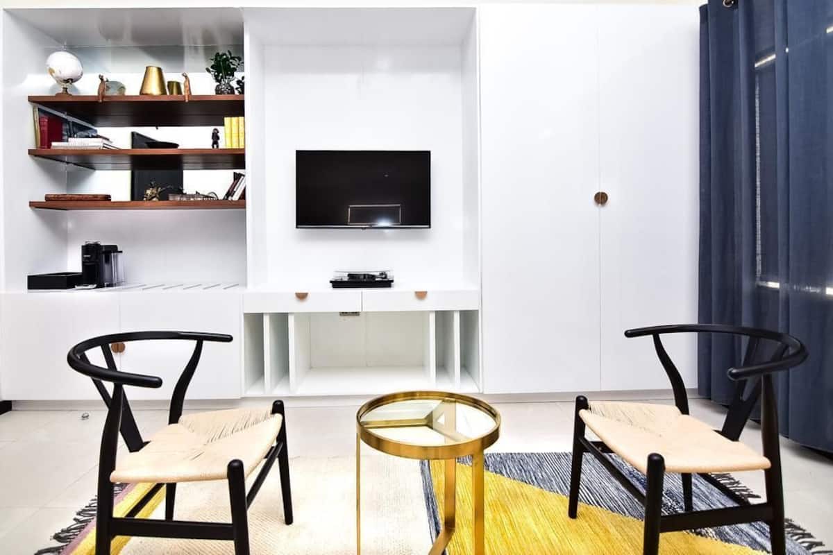 Uma sala de estar moderna com uma TV de tela plana na parede, duas cadeiras pretas com assentos trançados, uma pequena mesa de centro redonda e uma estante com diversos objetos.