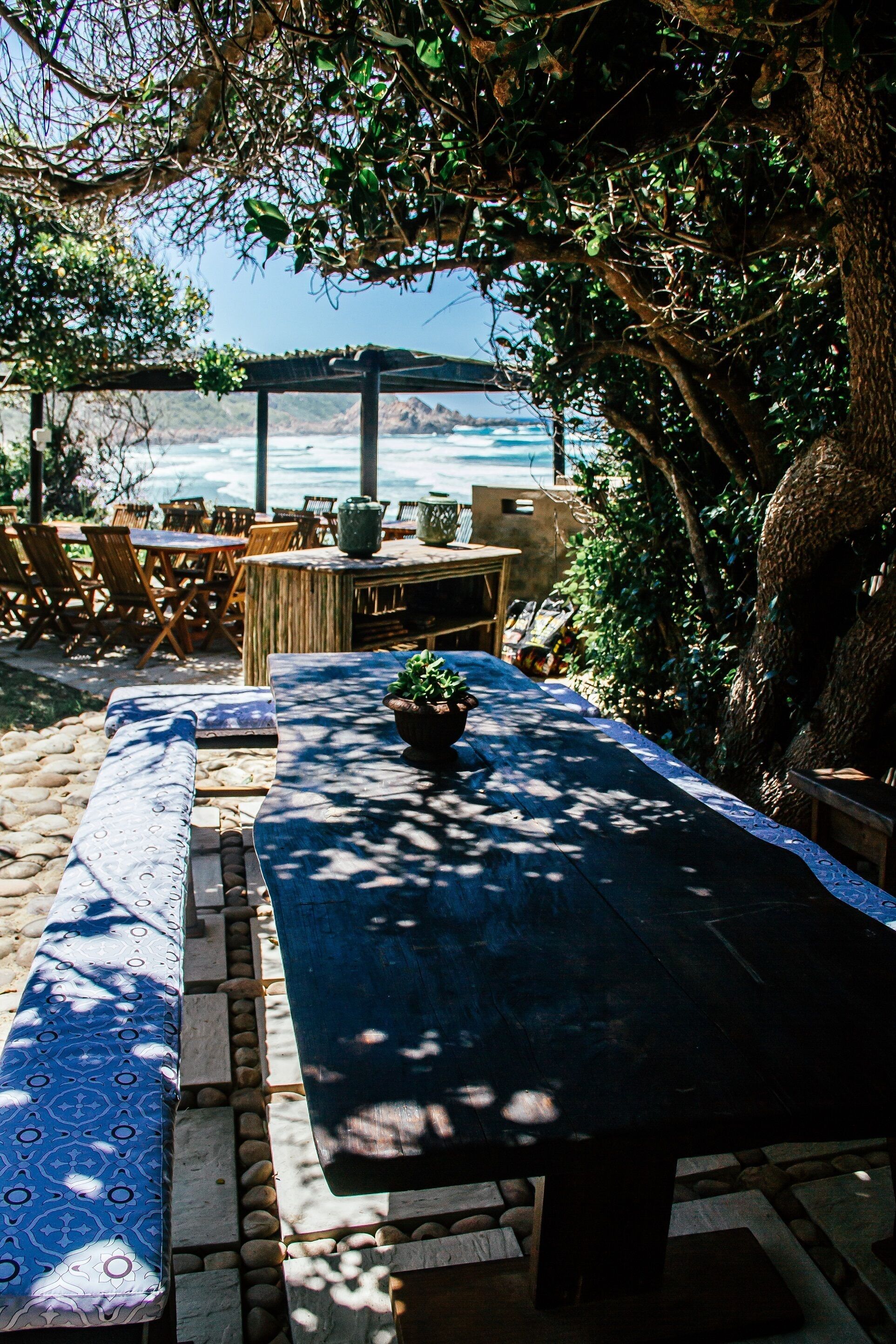 An outdoor seating area with a long table, benches, and chairs, overlooking a beach.