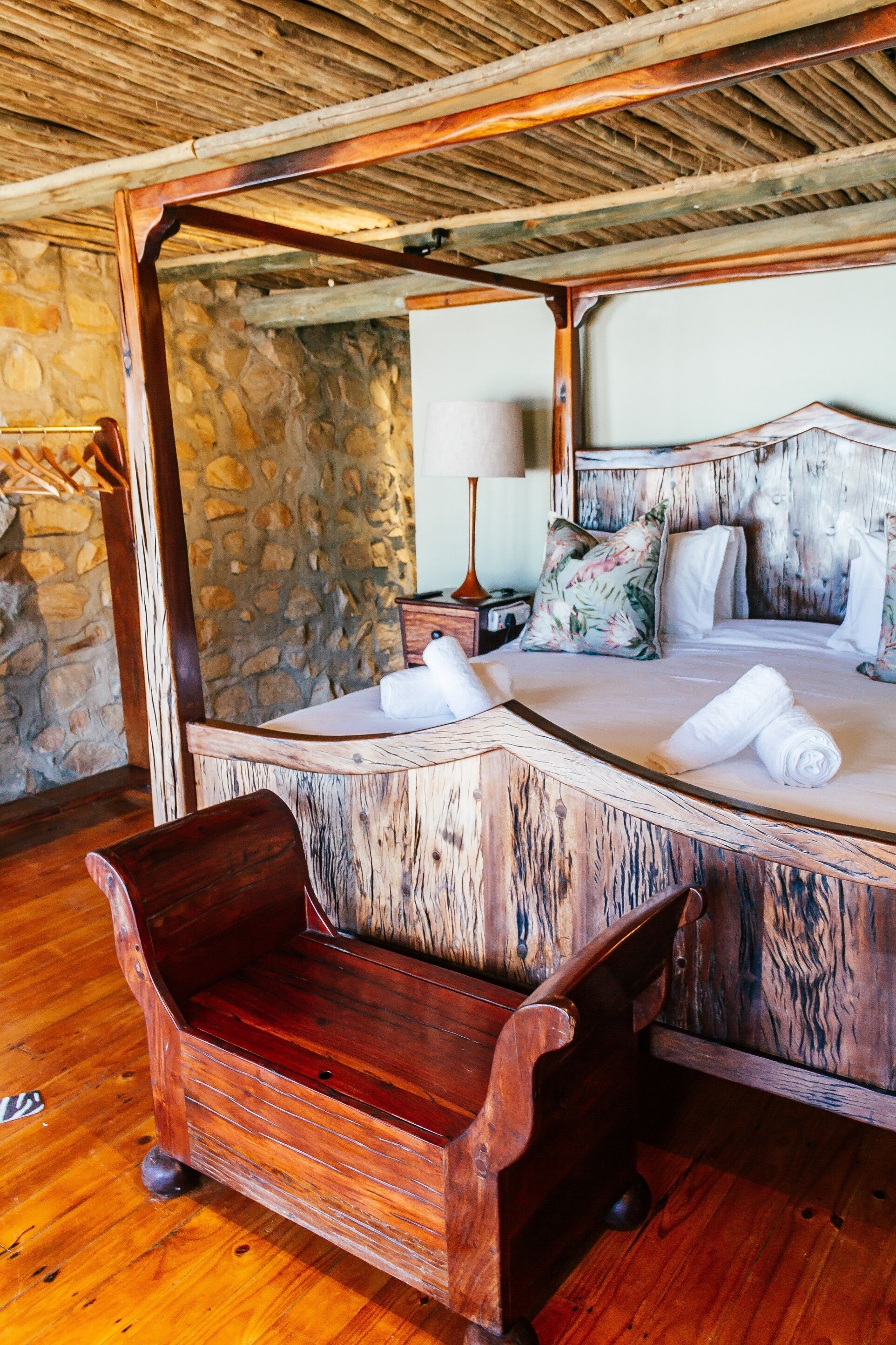 Una cama con estructura de madera, un fondo de pared de piedra, una lámpara de noche y un banco de madera.