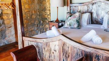 Una cama con estructura de madera, un fondo de pared de piedra, una lámpara de noche y un banco de madera.