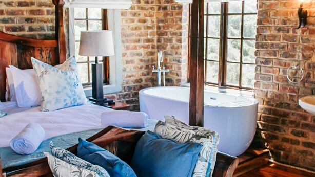 A rustic room with a wooden bed, a wooden bench, a large window, and a brick wall. - Buccara Noetzie - Lindsay Castle (Knysna)