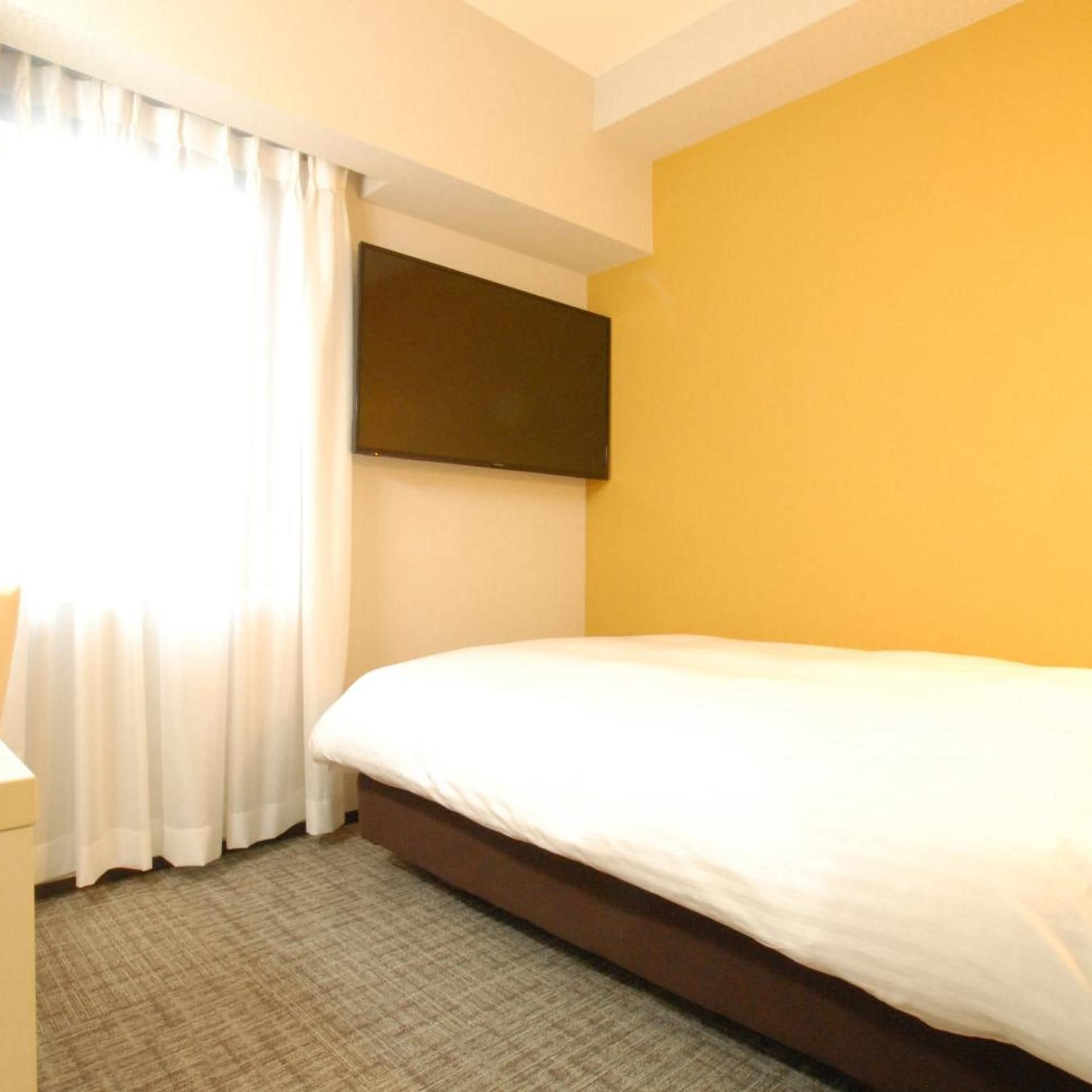 A hotel room with a bed, a flat-screen TV mounted on the wall, and a window with curtains.